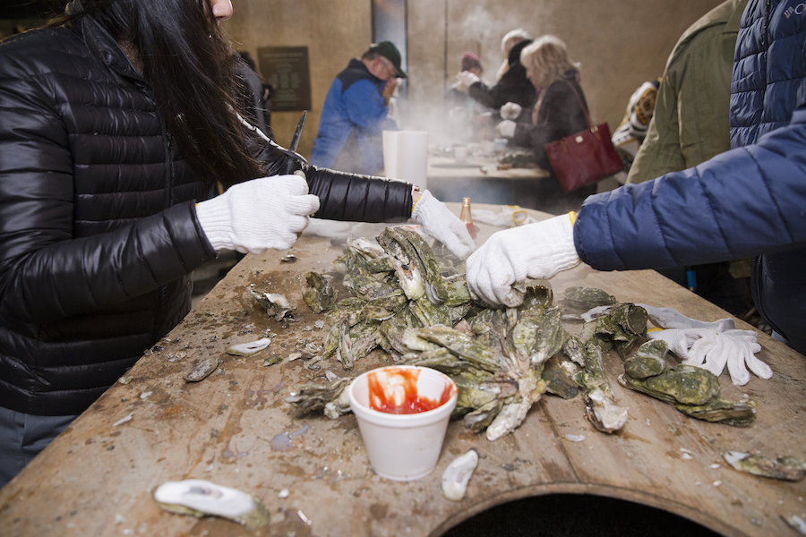 people in white gloves shuck fresh steamed oysters at a table outside