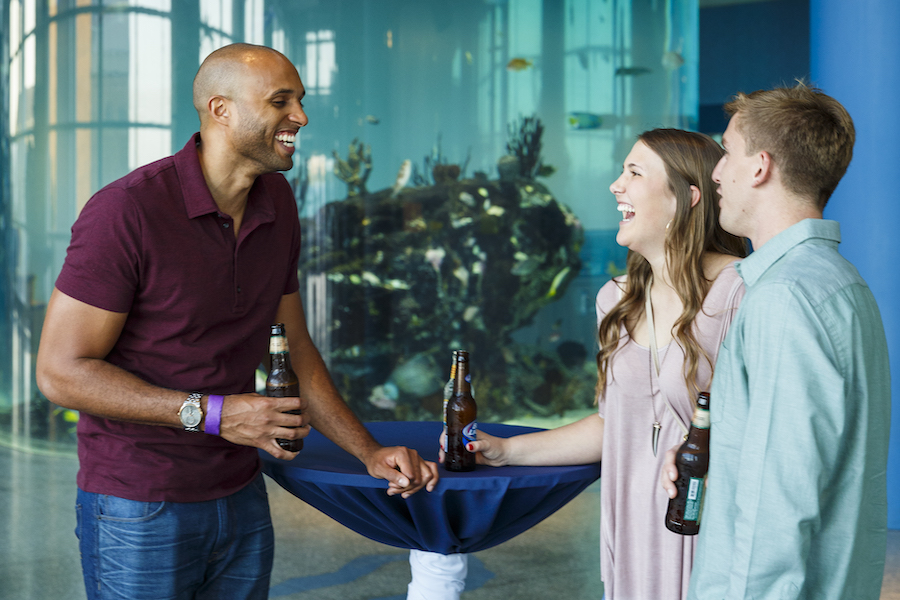 Two men and a woman stand at a cocktail table in front of Carolina Seas at South Carolina Aquarium. They laugh and talk as they drink from beer bottles.