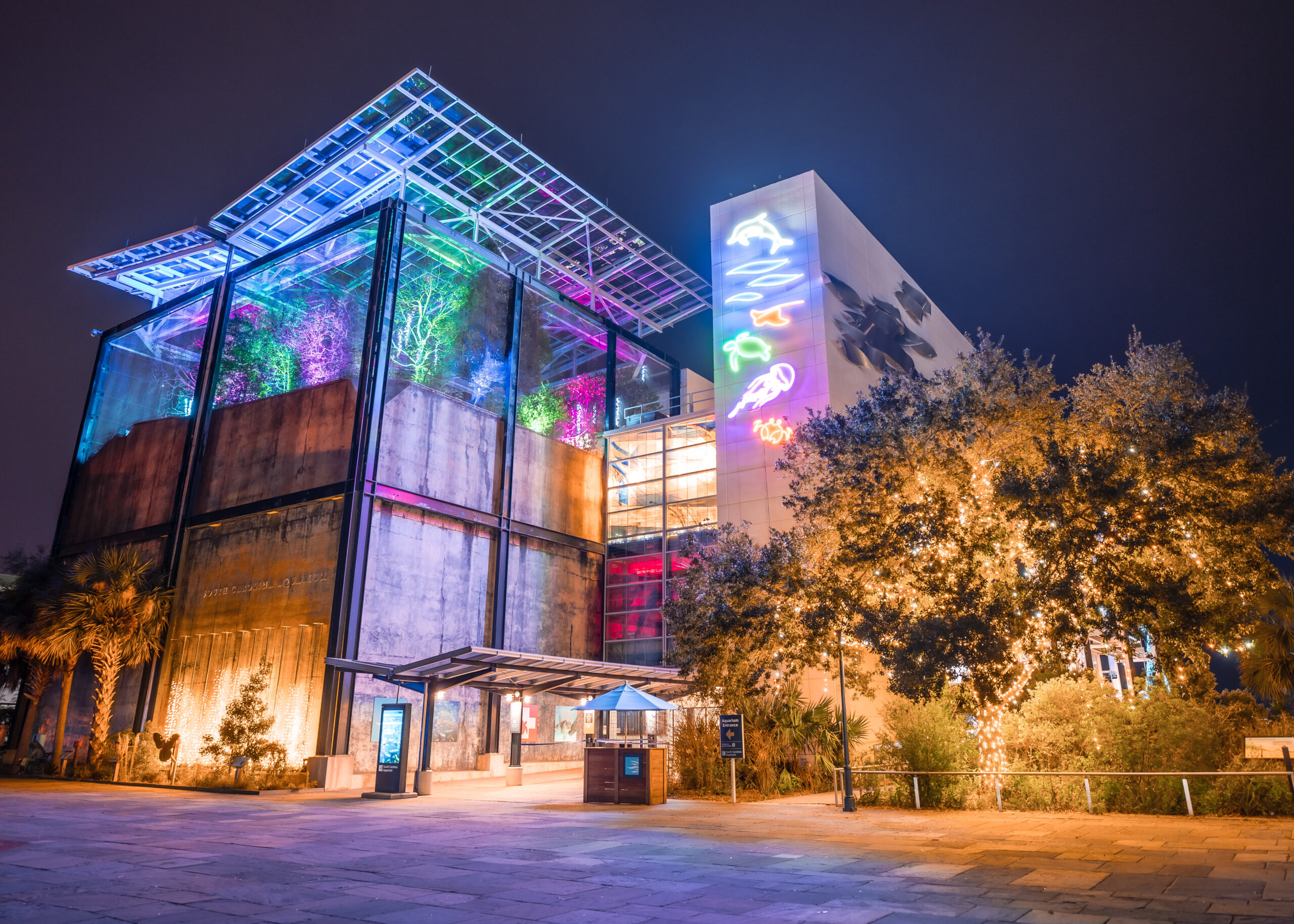 The exterior of the South Carolina Aquarium is shown at night, lit up by colorful blue, green and pink lights at the top, neon lights in animal shapes and gold glowing lights at the bottom and in surrounding trees.
